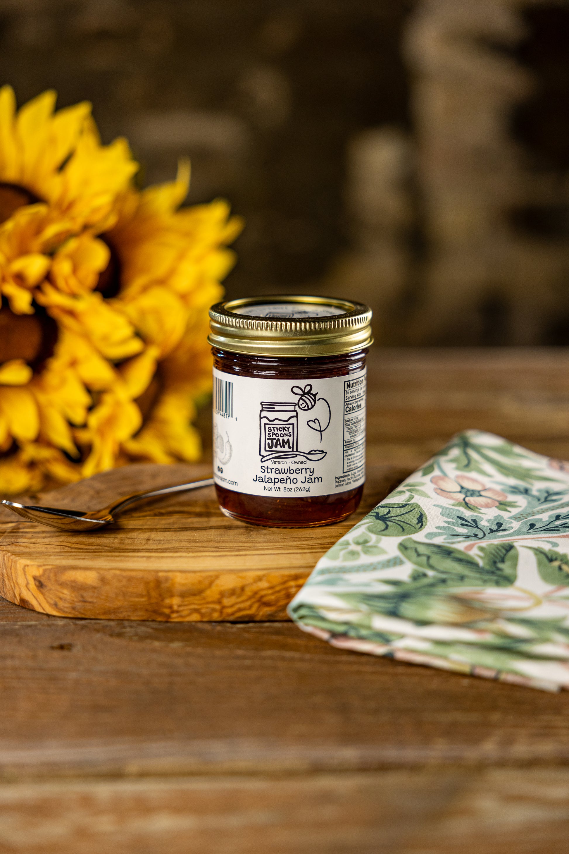 Jar of Sticky Spoons Strawberry Jalapeno jam on charcuterie board with spoon and napkin. Sunflower in background.